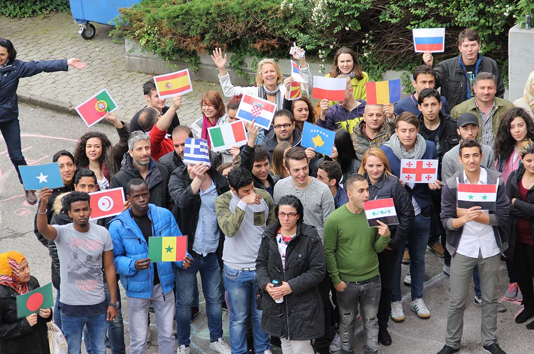 Gruppierung von Menschen im Innenhof, die Flaggen verschiedener Länder halten, darunter Bangladesch, Senegal, Griechenland, Italien und Syrien.